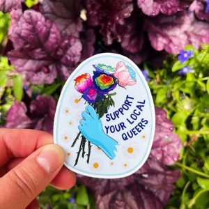 May include: A blue and white sticker with a rainbow rose bouquet and the text "Support Your Local Queers".