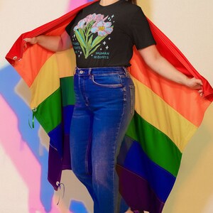 Puede incluir: Una persona lleva una camiseta negra con un dise&ntilde;o floral y el texto "Trans Rights Are Human Rights". Est&aacute; sosteniendo una bandera arco&iacute;ris del orgullo.