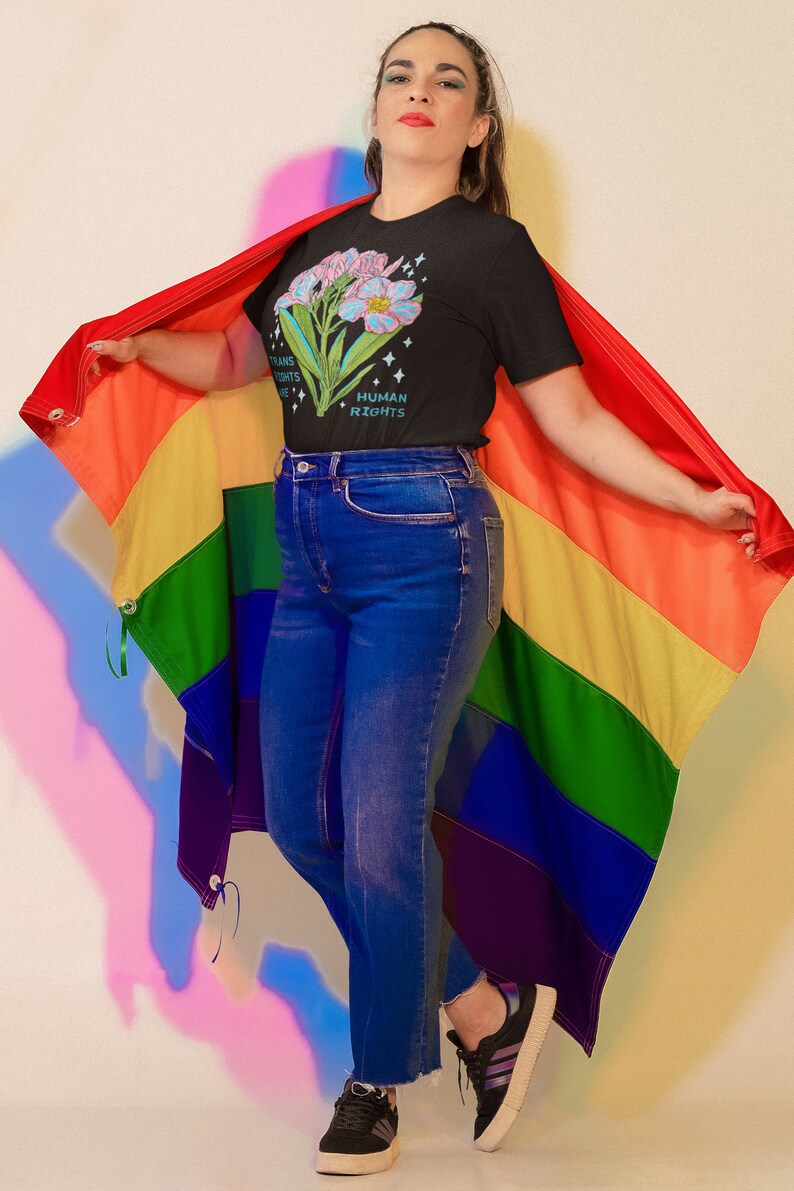 Puede incluir: Una persona lleva una camiseta negra con un dise&ntilde;o floral y el texto "Trans Rights Are Human Rights". Est&aacute; sosteniendo una bandera arco&iacute;ris del orgullo.