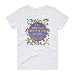 May include: White t-shirt with a quote from Audre Lorde in a blue circle with a floral border. The quote reads "I am deliberate and afraid of nothing."