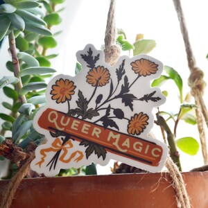 Puede incluir: Una pegatina blanca con un ramo de flores naranjas y hojas verdes. La pegatina tiene una pancarta que dice "Queer Magic" en texto amarillo sobre fondo rojo.