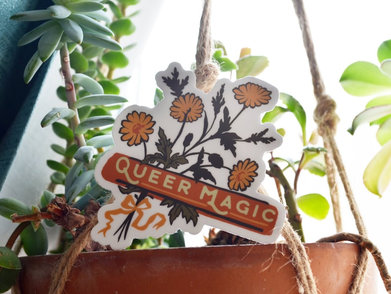 Puede incluir: Una pegatina blanca con un ramo de flores naranjas y hojas verdes. La pegatina tiene una pancarta que dice "Queer Magic" en texto amarillo sobre fondo rojo.
