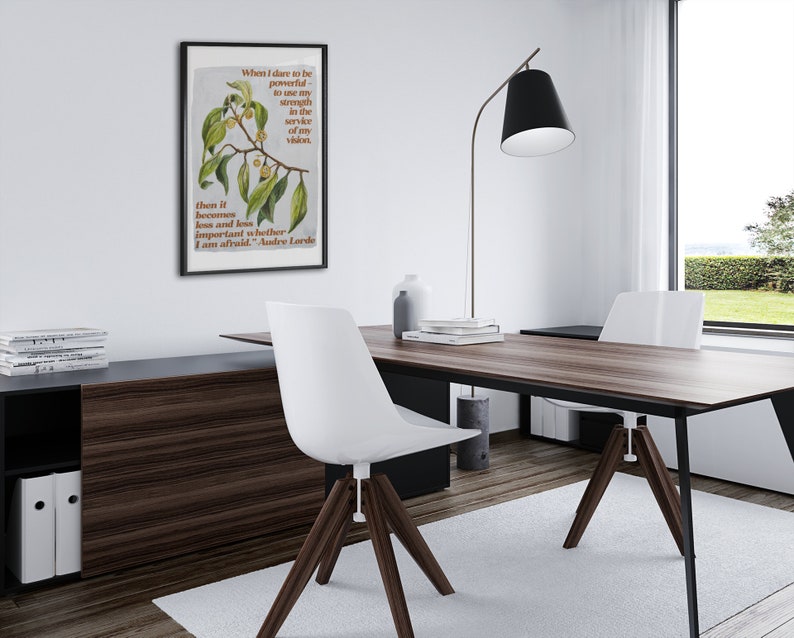K&ouml;nnte beinhalten: Ein schwarzer gerahmter Poster mit einem Zitat von Audre Lorde &uuml;ber die Verwendung von St&auml;rke im Dienste seiner Vision. Der Poster h&auml;ngt an einer wei&szlig;en Wand in einem modernen B&uuml;ro mit einem Holztisch, wei&szlig;en St&uuml;hlen und einer schwarzen Stehlampe.