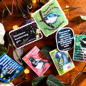 K&ouml;nnte beinhalten: Eine Sammlung farbenfroher Aufkleber mit verschiedenen Designs und Zitaten. Ein Aufkleber zeigt einen blauen Vogel, ein anderer sagt "Obstinate headstrong girl", und andere haben Illustrationen von Tieren und S&auml;tzen wie "It's always spooky season."