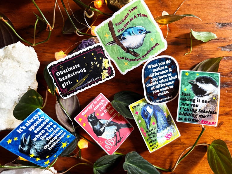 K&ouml;nnte beinhalten: Eine Sammlung farbenfroher Aufkleber mit verschiedenen Designs und Zitaten. Ein Aufkleber zeigt einen blauen Vogel, ein anderer sagt "Obstinate headstrong girl", und andere haben Illustrationen von Tieren und S&auml;tzen wie "It's always spooky season."
