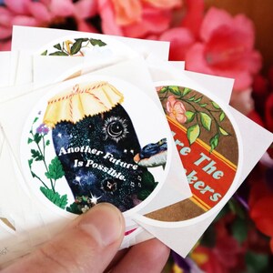 May include: A stack of round stickers with colourful illustrations and text. One sticker features a jar with a starry sky inside and the text "Another Future Is Possible". Another sticker features a brown background with green leaves and the text "We Are The Makers".