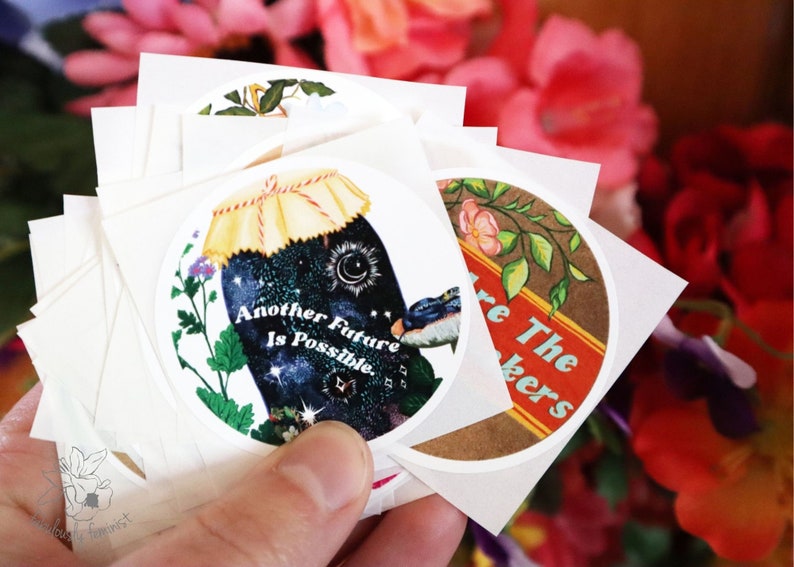 May include: A stack of round stickers with colourful illustrations and text. One sticker features a jar with a starry sky inside and the text "Another Future Is Possible". Another sticker features a brown background with green leaves and the text "We Are The Makers".