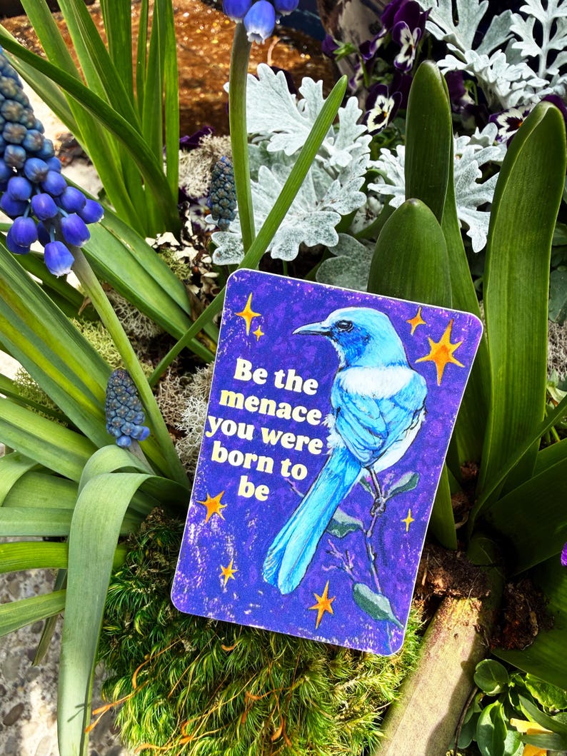 May include: A rectangular sticker with a blue bird illustration and the text "Be the menace you were born to be" on a purple background. The sticker is surrounded by green plants and blue flowers.