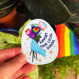 May include: A sticker with a rainbow rose bouquet, a blue gloved hand holding the stems, and the text "Support Your Local Queers" in white lettering on a white background.