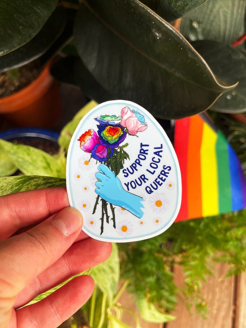 May include: A sticker with a rainbow rose bouquet, a blue gloved hand holding the stems, and the text "Support Your Local Queers" in white lettering on a white background.