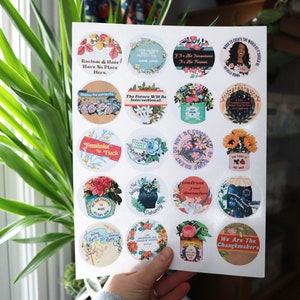 May include: A sheet of 18 round stickers featuring various feminist and social justice slogans and illustrations. The stickers include floral designs, a black cat, and a woman with an afro. Some of the slogans include "Racism & Hate Have No Place Here", "Feminist AF", "The Future Will Be Intersectional", and "We Are The Changemakers".