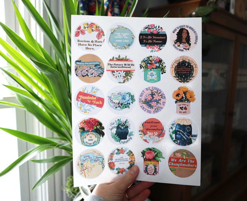 May include: A sheet of 18 round stickers featuring various feminist and social justice slogans and illustrations. The stickers include floral designs, a black cat, and a woman with an afro. Some of the slogans include "Racism & Hate Have No Place Here", "Feminist AF", "The Future Will Be Intersectional", and "We Are The Changemakers".