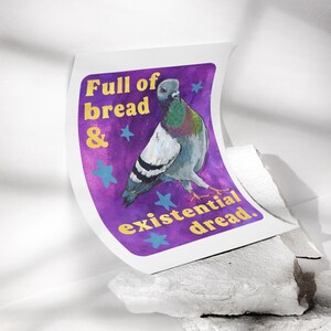 K&ouml;nnte beinhalten: Ein rechteckiger Kunstdruck mit einer grauen Taube mit schillernden Halsfedern vor einem violetten Hintergrund. Der Text lautet Full of bread & existential dread in gelben Buchstaben, akzentuiert durch hellblaue Sterne im Design.
