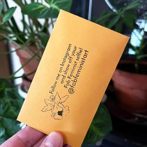 May include: A mustard-yellow card with black text and a floral illustration. The text reads "Follow me on Instagram and show off your Fab Feminist selfie! @fabfeministart".