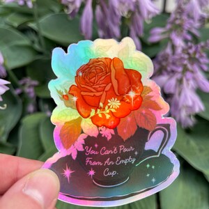 May include: A holographic sticker with a rose in a teacup. The sticker has a rainbow iridescent finish and features the text "You Can't Pour From An Empty Cup."