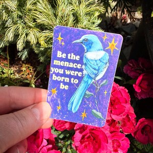 May include: A rectangular sticker with a blue bird illustration on a purple background. The sticker has the text "Be the menace you were born to be" and yellow star accents. The sticker is held in front of a background of red flowers and green foliage.
