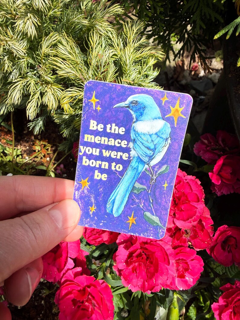 May include: A rectangular sticker with a blue bird illustration on a purple background. The sticker has the text "Be the menace you were born to be" and yellow star accents. The sticker is held in front of a background of red flowers and green foliage.