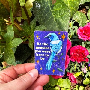May include: A rectangular sticker with a blue bird illustration and the text "Be the menace you were born to be." The sticker has a purple background with yellow star accents. The sticker is held in front of a background of green leaves and pink flowers.