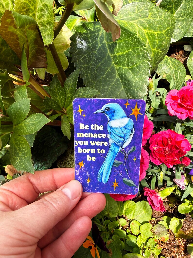 May include: A rectangular sticker with a blue bird illustration and the text "Be the menace you were born to be." The sticker has a purple background with yellow star accents. The sticker is held in front of a background of green leaves and pink flowers.