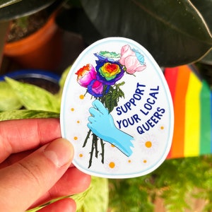May include: A sticker with a blue background and a hand holding a bouquet of rainbow roses. The text on the sticker reads "Support Your Local Queers".