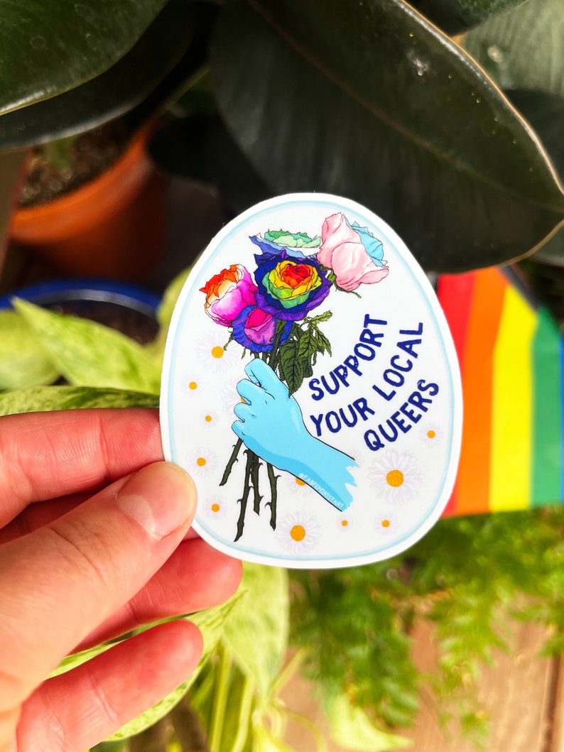 May include: A sticker with a blue background and a hand holding a bouquet of rainbow roses. The text on the sticker reads "Support Your Local Queers".