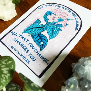 Peut inclure: Une impression blanche avec une illustration de fleur rose et bleue et le texte "All that you touch, you change. All that you change, changes you. Octavia Butler."