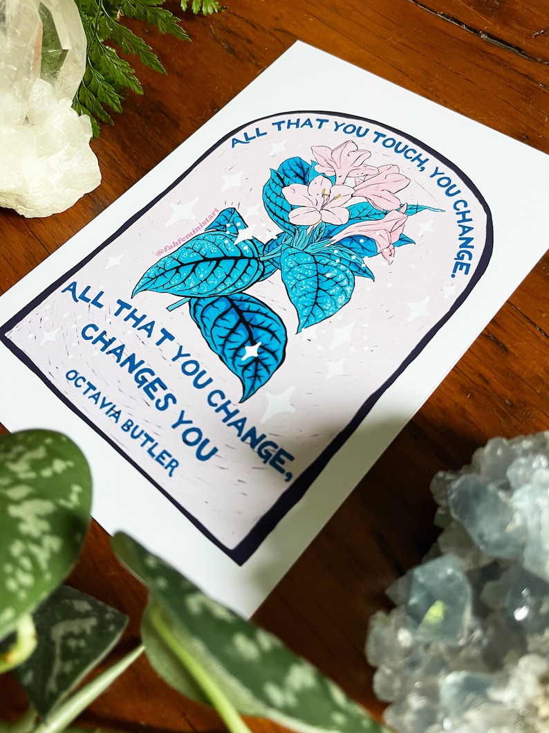 Peut inclure: Une impression blanche avec une illustration de fleur rose et bleue et le texte "All that you touch, you change. All that you change, changes you. Octavia Butler."