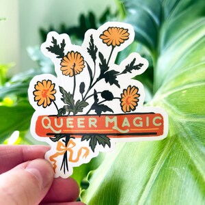 Puede incluir: Una pegatina con un ramo de flores naranjas y hojas verdes. La pegatina tiene una pancarta roja con el texto "QUEER MAGIC" en letras blancas.