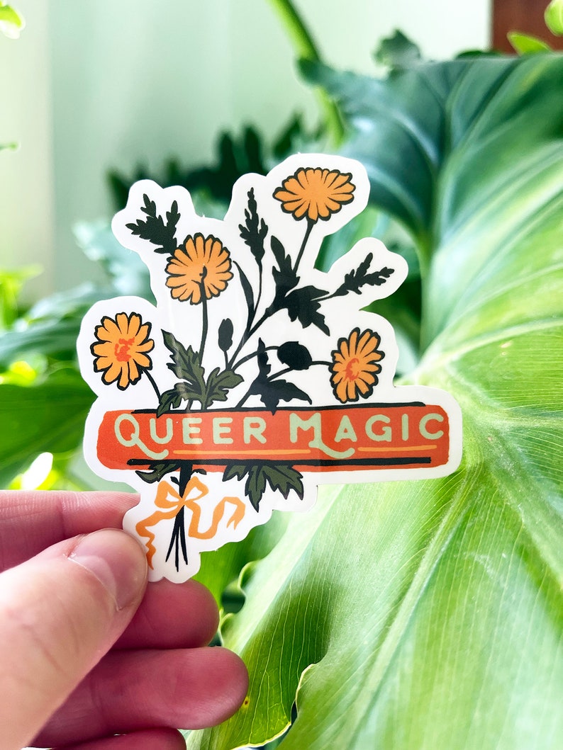 Puede incluir: Una pegatina con un ramo de flores naranjas y hojas verdes. La pegatina tiene una pancarta roja con el texto "QUEER MAGIC" en letras blancas.