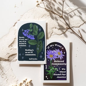 May include: Two illustrated cards with a dark blue background and white text. The first card features a purple flower with green leaves and the text "Rarely, if ever, are any of us healed in isolation. Healing is an act of communion. bell hooks." The second card features a purple flower with green leaves and the text "If We Want A Beloved Community We Must Stand For Justice."