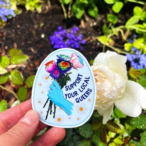 May include: A blue and white sticker with a rainbow rose bouquet and the text "Support Your Local Queers".