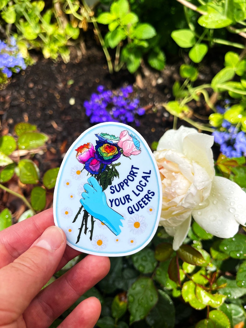 May include: A blue and white sticker with a rainbow rose bouquet and the text "Support Your Local Queers".