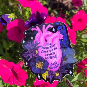 Puede incluir: Una pegatina en forma de coraz&oacute;n rosa con fondo negro y detalles florales. La pegatina presenta la cita "El coraz&oacute;n de la justicia es decir la verdad" de bell hooks.