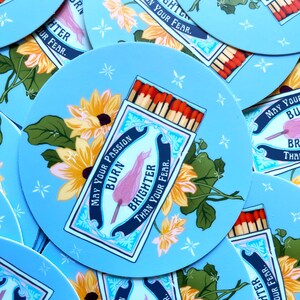 Pu&ograve; includere: Un adesivo rotondo con sfondo blu e stelle bianche. L'adesivo presenta una scatola di fiammiferi con un fiammifero rosa che dice "Che la tua passione bruci pi&ugrave; forte della tua paura." La scatola di fiammiferi &egrave; circondata da fiori gialli e foglie verdi.