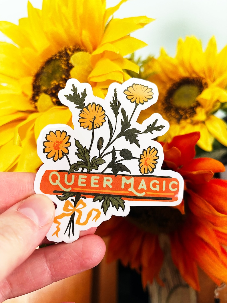 Puede incluir: Una pegatina con un ramo de flores amarillas y una pancarta que dice "Queer Magic". La pegatina se sostiene en una mano sobre un fondo de girasoles.
