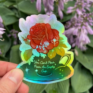 May include: A holographic sticker with a green teacup and a red rose. The teacup has a white rim and a handle. The text on the sticker reads "You Can't Pour From An Empty Cup."