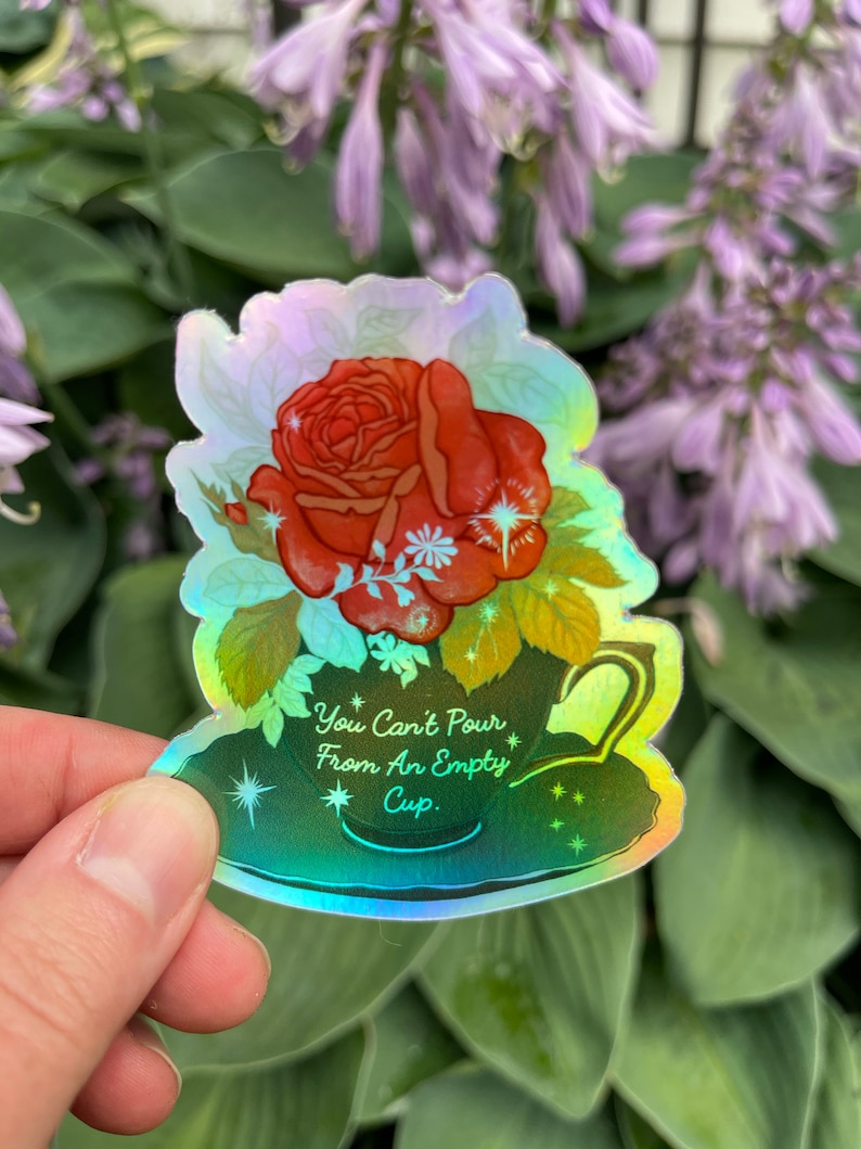 May include: A holographic sticker with a green teacup and a red rose. The teacup has a white rim and a handle. The text on the sticker reads "You Can't Pour From An Empty Cup."