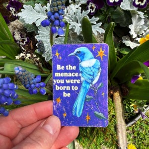 May include: A rectangular card with a blue bird illustration and the text "Be the menace you were born to be." The card is held in front of a background of purple flowers and greenery.
