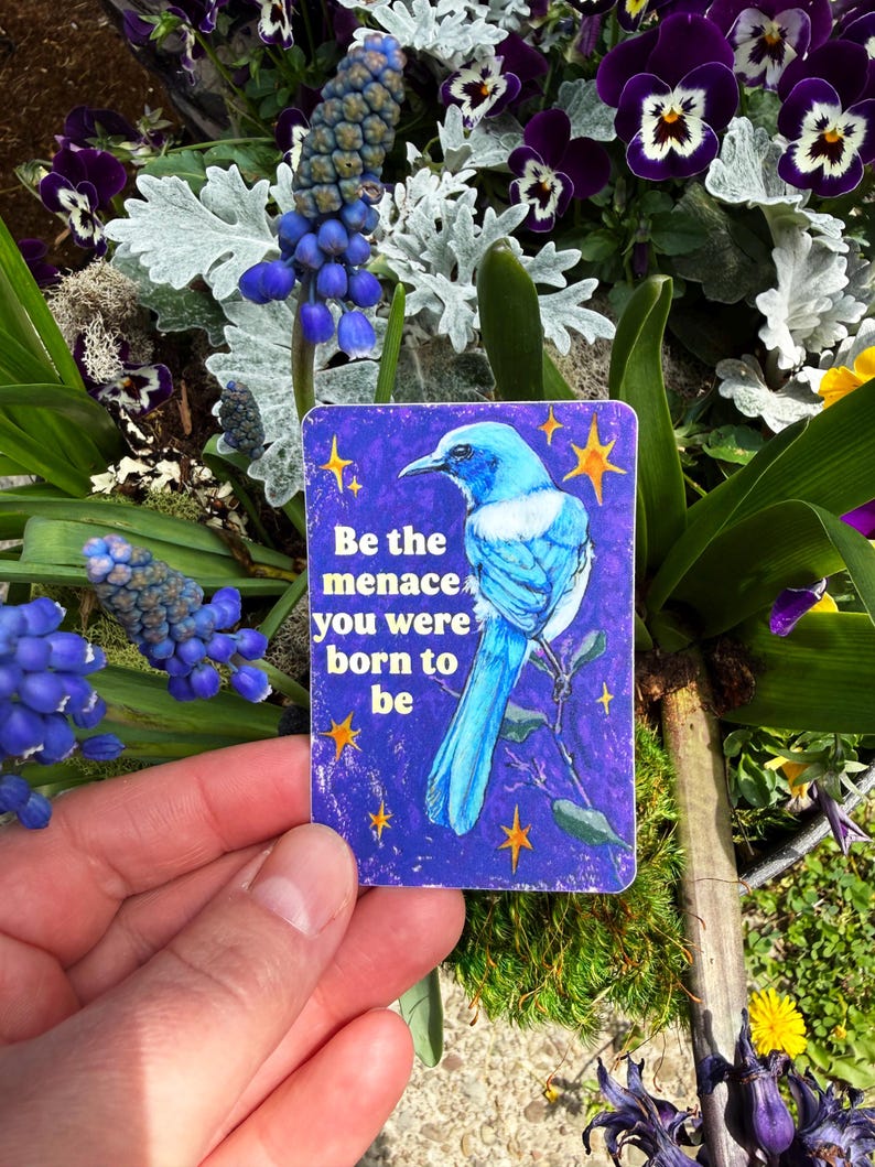 May include: A rectangular card with a blue bird illustration and the text "Be the menace you were born to be." The card is held in front of a background of purple flowers and greenery.