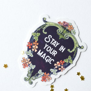 May include: A sticker with a dark blue background and white text that reads "Stay in your magic". The sticker is surrounded by a green and gold floral design.