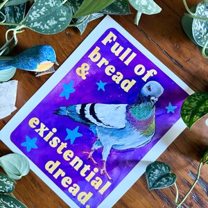 K&ouml;nnte beinhalten: Ein rechteckiger Kunstdruck mit einem Taubenmotiv vor einem leuchtend violetten und blauen Aquarellhintergrund. Der Text Full of bread & existential dread steht in gelben Buchstaben, umgeben von kleinen blauen Sternen.