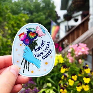 May include: A sticker with a blue gloved hand holding a bouquet of rainbow roses. The sticker has a white background with the text "Support Your Local Queers" in blue and white lettering.