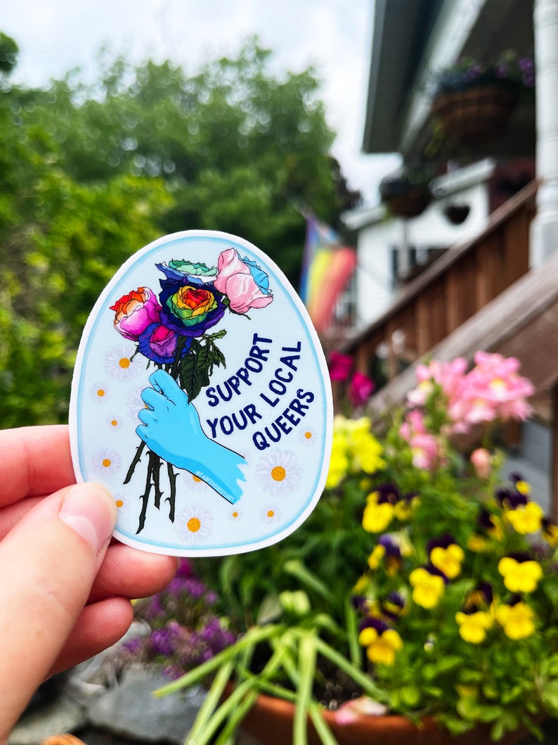 May include: A sticker with a blue gloved hand holding a bouquet of rainbow roses. The sticker has a white background with the text "Support Your Local Queers" in blue and white lettering.