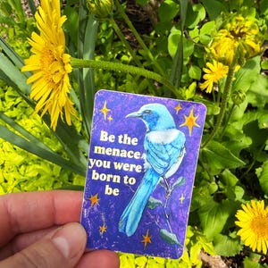 May include: A rectangular card with a blue bird illustration and the text "Be the menace you were born to be" is held in front of yellow flowers and green foliage. The card has a purple background with yellow star accents.