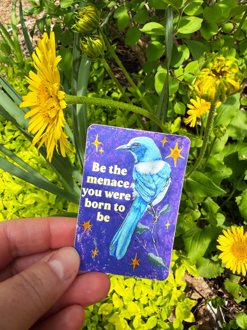 May include: A rectangular card with a blue bird illustration and the text "Be the menace you were born to be" is held in front of yellow flowers and green foliage. The card has a purple background with yellow star accents.