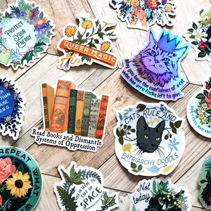 Puede incluir: Una colecci&oacute;n de pegatinas coloridas con varios temas feministas y LGBTQ+. Las pegatinas presentan dise&ntilde;os florales, gatos, libros y texto como "Protege tu energ&iacute;a", "Magia queer", "Lee libros y desmantela los sistemas de opresi&oacute;n", "Todos los cuerpos son buenos cuerpos" y "Los gatos mandan y el patriarcado babea".