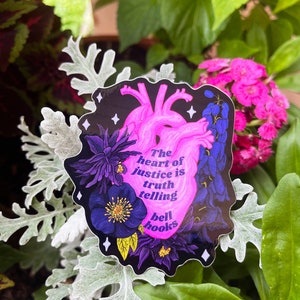 Puede incluir: Una pegatina con una ilustraci&oacute;n de coraz&oacute;n rosa y el texto "The heart of justice is truth telling bell hooks" sobre un fondo negro con flores moradas y azules.