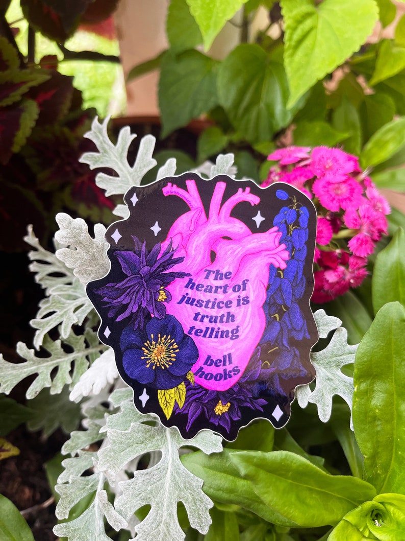 Puede incluir: Una pegatina con una ilustraci&oacute;n de coraz&oacute;n rosa y el texto "The heart of justice is truth telling bell hooks" sobre un fondo negro con flores moradas y azules.