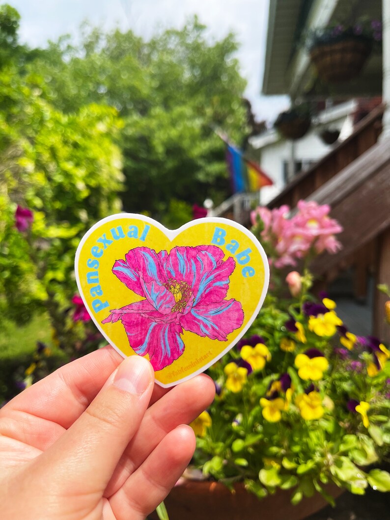 May include: A yellow heart-shaped sticker with a pink and blue flower and the text "pansexual babe".