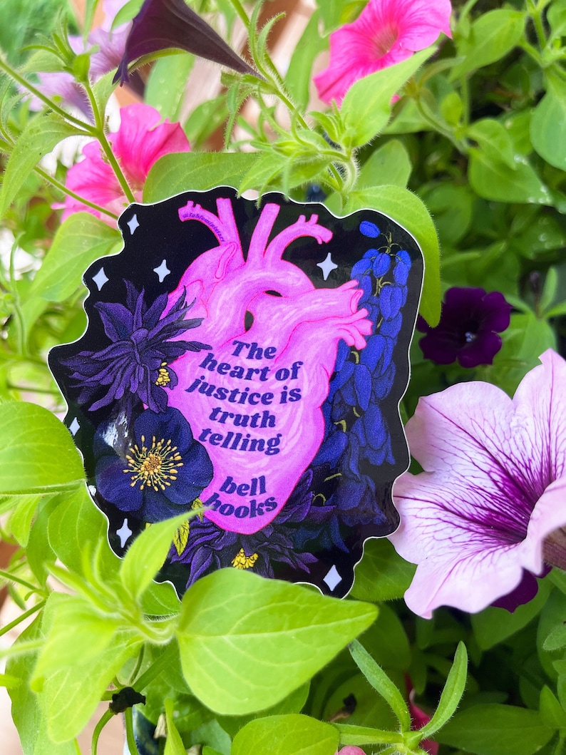 Puede incluir: Un coraz&oacute;n rosa con el texto "The heart of justice is truth telling bell hooks" sobre un fondo negro con flores moradas y azules.